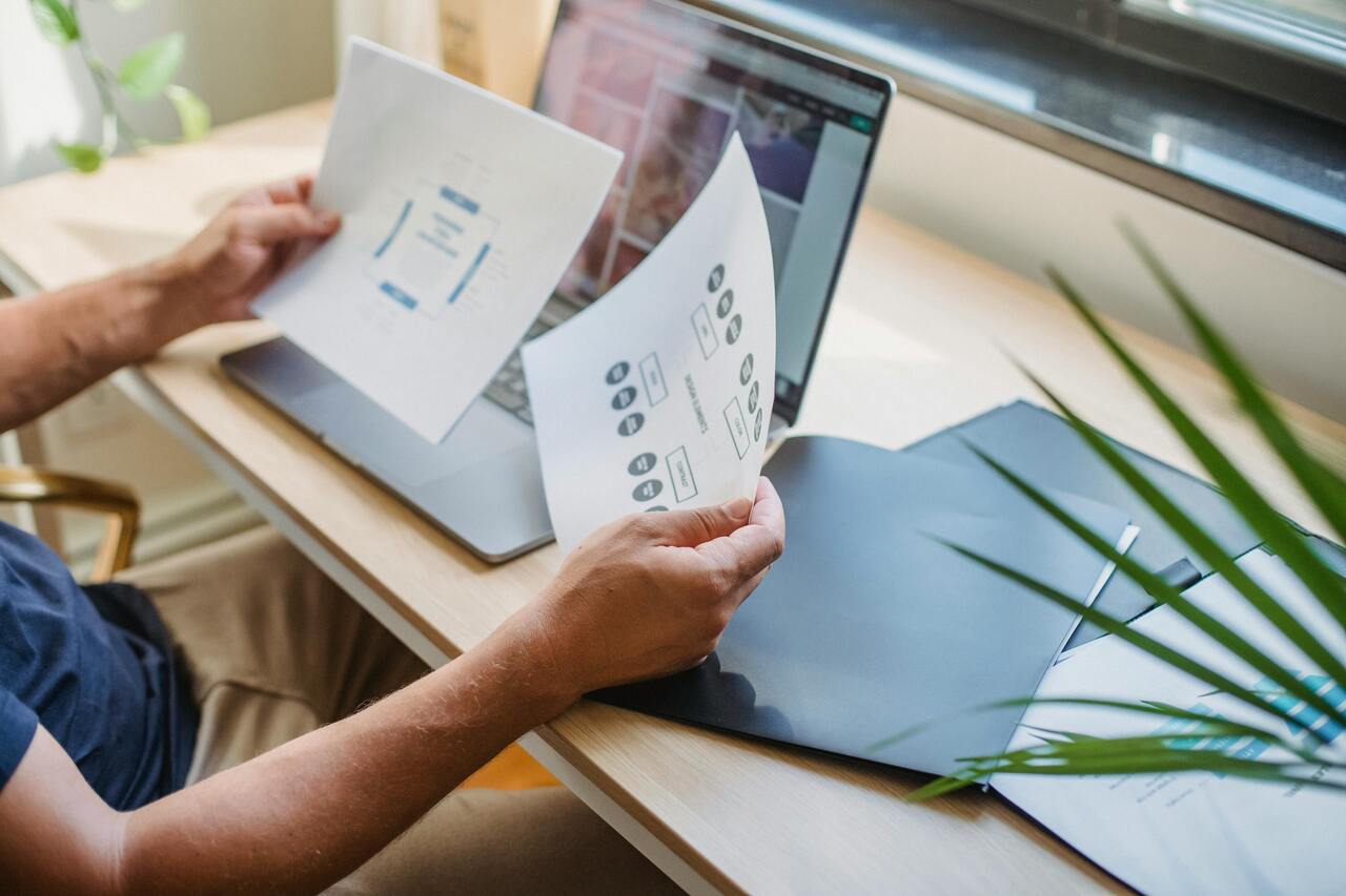 Persoon bekijkt geprinte schema’s en documenten aan een bureau met laptop tijdens het werken aan website- of strategieontwerp.